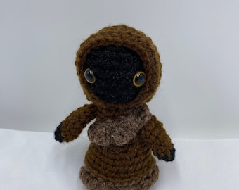 Puede incluir: Un juguete de peluche de color marrón tejido a crochet con ojos negros y una túnica con capucha marrón. El juguete está de pie sobre una superficie blanca.