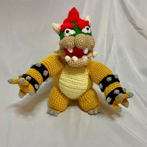 Könnte beinhalten: Handgefertigtes Bowser-Plüschtier mit gelbem Körper, grün-rotem Kopf und schwarzen Armbändern mit Stacheln. Die Häkelfigur hat ein detailliertes Gesicht mit weißen Zähnen und einem roten Mund sowie grauen Krallen.