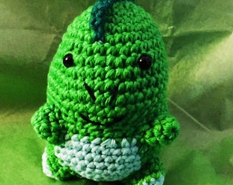 Puede incluir: Juguete de dinosaurio de crochet verde con una cresta azul y ojos negros. El dinosaurio tiene una expresión sonriente y una barriga azul claro.