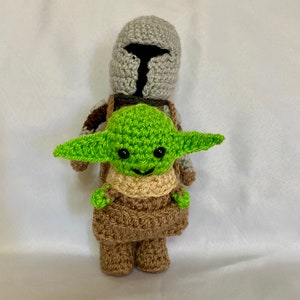 Puede incluir: Una figura de Mandalorian hecha a crochet que lleva una figura de Baby Yoda hecha a crochet. El Mandaloriano lleva un casco gris y una armadura marr&oacute;n. Baby Yoda lleva una t&uacute;nica verde y tiene ojos grandes y negros.