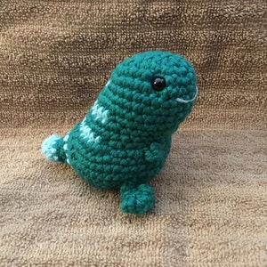 May include: A handmade teal crochet seal plushie with white and light blue accents. The seal has black eyes and a white stitched smile. The plushie has flippers and a tail. The background is a textured brown surface.