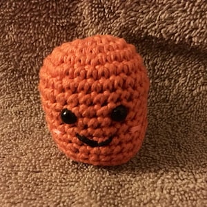 May include: A small crocheted orange toy with a smiling face. The toy has black eyes and a black mouth.