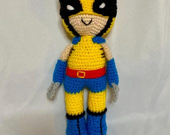 Op de afbeelding: Een gehaakt pluche speelgoed van Wolverine uit de X-Men. Het speelgoed is geel met zwarte klauwen, een zwart masker en een rode riem. Het speelgoed draagt een blauwe broek en laarzen.