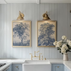 Pode incluir: Duas impressões emolduradas de árvores azuis e brancas penduradas acima de uma pia branca de fazenda com uma torneira de latão. A cozinha apresenta armários e paredes azuis claros, arandelas douradas e um vaso de flores brancas.