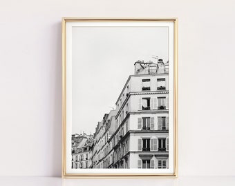May include: Black and white framed print of Parisian architecture. The image features a row of buildings with multiple stories, windows, and balconies. The buildings are in a classic style, with sloped roofs and chimneys. The print is in a gold frame.