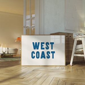 May include: A framed poster with the text "WEST COAST" in blue letters on a white background. The poster is leaning against a wooden cabinet in a room with a white wall and wooden floors.