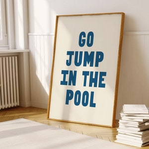 May include: A framed poster with a white background and blue text that reads "GO JUMP IN THE POOL". The poster is hanging on a white wall in a room with a wooden floor.
