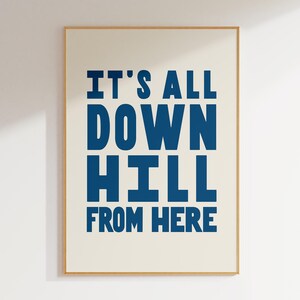 May include: A framed print featuring the phrase "IT'S ALL DOWN HILL FROM HERE" in dark blue, bold text on a cream background. The print is encased in a light brown wooden frame and hangs on a white wall.