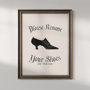 Pode incluir: Impressão emoldurada com uma ilustração vintage de um sapato preto e o texto "Please Remove Your Shoes OR PERISH" em escrita elegante. A obra de arte está sobre um fundo creme, emoldurada em uma moldura de madeira escura.