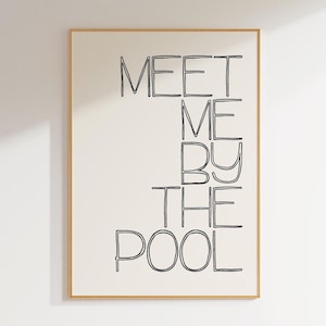 May include: A framed art print with the words "MEET ME BY THE POOL" in black, handwritten-style font. The text is centered on a cream-colored background, encased in a thin, light brown wooden frame. The artwork is displayed against a white wall.