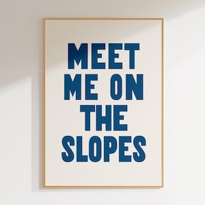 Könnte beinhalten: Ein gerahmter Kunstdruck mit dem Text "MEET ME ON THE SLOPES" in fetten, dunkelblauen Buchstaben. Der Text ist auf einem cremefarbenen Hintergrund zentriert und von einem dünnen, hellbraunen Rahmen umgeben. Das Kunstwerk hängt an einer weißen Wand.