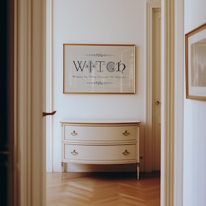 Puede incluir: Un estampado enmarcado con la palabra "WITCH" en una fuente decorativa, con el texto "Women In Total Control Of Herself" debajo. El estampado cuelga sobre una c&oacute;moda blanca y dorada con dos cajones. La habitaci&oacute;n tiene una paleta de colores neutros.