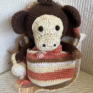 May include: A crocheted monkey backpack with a striped fabric lining. The monkey has brown fur, black eyes, and a beige face. The backpack has a long strap and is made with beige, brown, and orange yarn.