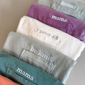 May include: A stack of eight folded t-shirts in various colors, each with a different embroidered phrase on the pocket. The phrases include "mama", "be kind", "I Peter 4:8", and "mama".