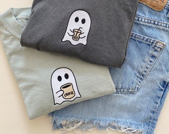 May include: Two gray and light blue t-shirts with embroidered ghost designs. The ghost on the gray shirt is holding a boba tea, and the ghost on the light blue shirt is holding a coffee cup. The shirts are folded on top of a pair of light blue denim shorts.