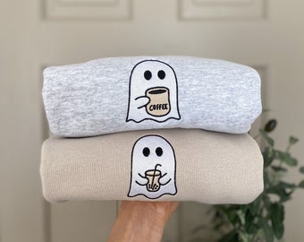 May include: Two sweatshirts stacked on top of each other. The top sweatshirt is gray and features a ghost holding a coffee cup with the word "COFFEE" on it. The bottom sweatshirt is beige and features a ghost holding a boba tea cup.