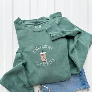 May include: A green sweatshirt with the text "Bring me an iced coffee" and an embroidered image of a cup of iced coffee.