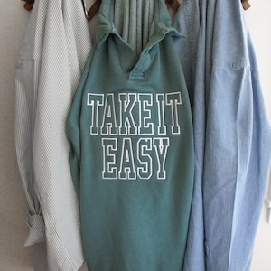 May include: A teal green hooded sweatshirt with the words "TAKE IT EASY" in white block letters hangs on a wooden coat rack, flanked by a striped shirt and a denim shirt.