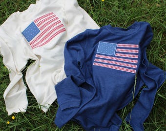 May include: Two long-sleeved shirts featuring American flag designs. One shirt is white with a red, white and blue gingham flag. The other shirt is blue with a red, white and blue gingham flag.
