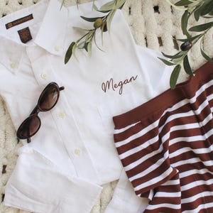 May include: A white button-down shirt with the name "Megan" embroidered on the pocket, paired with brown and white striped shorts and sunglasses. Olive branches with olives are placed on a textured surface.