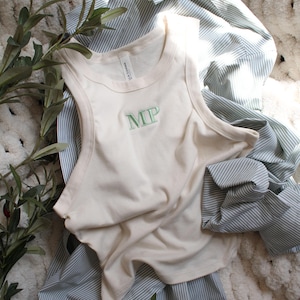 May include: Cream-colored tank top with the embroidered letters "MP" in green, layered over a blue and white striped button-down shirt. The clothing is arranged on a textured, off-white surface, with olive branches in the foreground.