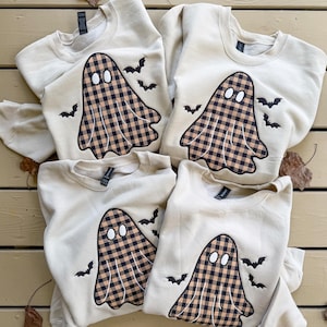 May include: Four cream-colored crewneck sweatshirts, each featuring an embroidered ghost with a brown and black buffalo plaid pattern. The ghost design includes two white oval eyes and is surrounded by several small, black embroidered flying bats.