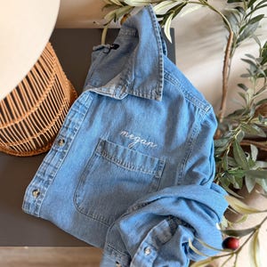 May include: A light blue denim button-down shirt featuring a classic collar, long sleeves with buttoned cuffs, and a single chest pocket. The name "megan" is embroidered in white cursive script above the pocket on the front of the garment.