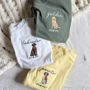 May include: Three t-shirts with embroidered dog designs. A white shirt features a brown Labrador, a yellow shirt has a Pointer, and a green shirt displays a Golden Retriever. Each shirt has the word "mom" embroidered below the dog.