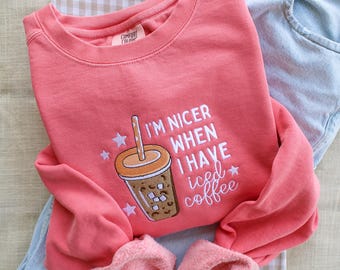 Op de afbeelding: Een roze sweatshirt met de tekst "Ik ben aardiger als ik ijskoffie heb" en een geborduurd beeld van een kopje ijskoffie.