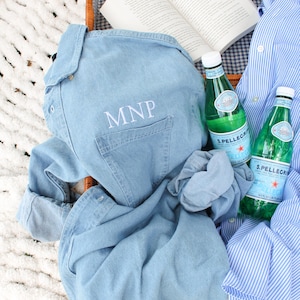 May include: A light blue denim shirt with the embroidered initials "MNP" on the pocket, alongside a blue and white striped shirt, two bottles of S.Pellegrino, and an open book inside a picnic basket.