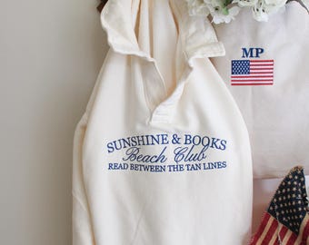 Könnte beinhalten: Ein weißer Kapuzenpullover mit einem blauen gestickten Logo, auf dem "Sunshine & Books Beach Club Read Between The Tan Lines" steht.