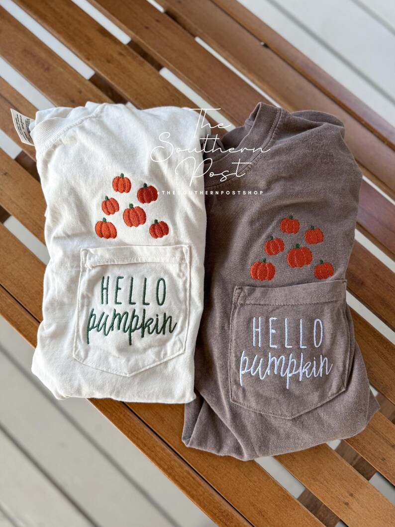 May include: Two t-shirts with a pocket on the front. The pocket has the text "Hello Pumpkin" embroidered on it. The t-shirts are in different colors, one is white and the other is brown. Both t-shirts have embroidered pumpkins on the pocket.