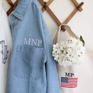 May include: A light blue denim shirt with a white embroidered pocket that says "MNP" hanging on a wooden rack next to a white canvas tote bag with a red, white, and blue American flag and the letters "MP" embroidered on it. The tote bag has a bouquet of white flowers.