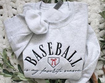 May include: A gray sweatshirt with the text "Baseball is my favorite season" in black embroidery. A red baseball diamond with a red bow is in the center of the text.