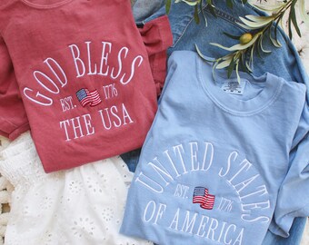 May include: Two long-sleeved shirts with patriotic embroidery. One is a dusty rose color with "GOD BLESS THE USA" and a flag. The other is light blue with "UNITED STATES OF AMERICA" and a flag. A denim jacket and white eyelet skirt are also visible.