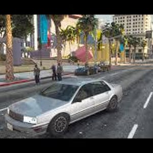 May include: A silver saloon car is driving on a multi-lane road. The car is in the foreground, with buildings and palm trees in the background. Several people are standing on the pavement.