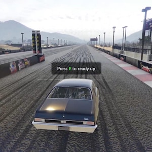 Puede incluir: Un coche clásico negro está posicionado en una pista de carreras de aceleración, con el texto "Press T to ready up" mostrado. La pista es de asfalto, con una cordillera al fondo. El velocímetro del coche muestra 1 mph.