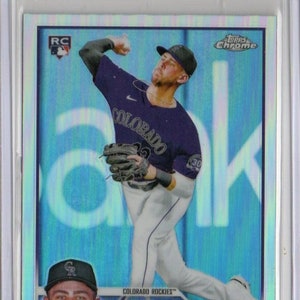 Puede incluir: Una tarjeta de béisbol con Nolan Jones, un jugador novato de los Colorado Rockies. La tarjeta es una tarjeta Topps Chrome con un fondo holográfico azul y plateado. La tarjeta presenta una foto de Jones lanzando, con el logotipo de los Colorado Rockies y "Nolan Jones" impreso en la tarjeta.