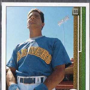 Puede incluir: Una tarjeta de béisbol con Omar Vizquel, un jugador de béisbol que lleva una camiseta azul de los Mariners con letras amarillas. La tarjeta tiene un borde verde, dorado y blanco con el texto "SS" y "M's" en ella.