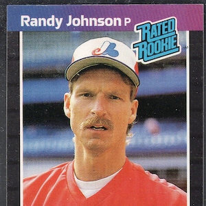 Puede incluir: Una tarjeta de béisbol con Randy Johnson, un lanzador, con el texto "Randy Johnson P" en la parte superior. La tarjeta incluye el logo "Rated Rookie" y el logo Donruss '89. Johnson lleva una camiseta roja y una gorra de béisbol.