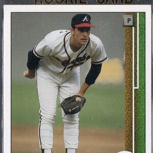 Puede incluir: Una tarjeta de béisbol con un jugador de béisbol que lleva un uniforme de béisbol blanco y rojo con una gorra de béisbol roja y blanca. El jugador está sosteniendo un guante de béisbol y está en posición de lanzar. La tarjeta tiene un borde dorado y el texto "Rookie" y "John Smoltz" en ella.