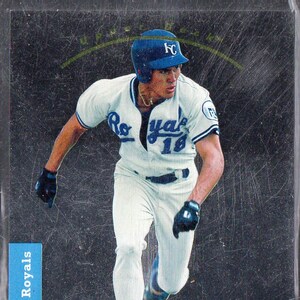 May include: A baseball card featuring a baseball player wearing a white jersey with the word "Royals" on the front, a blue cap with the letter "K" on it, and white trousers. The player is running with a baseball bat in his hand. The card has a black background with a gold border and the text "Kansas City Royals" in blue.