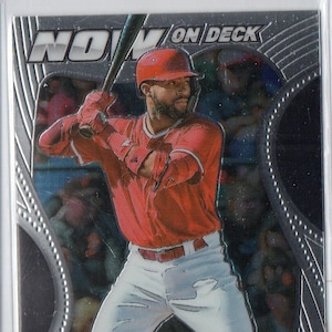 Peut inclure: Une carte de baseball argentée représentant un joueur de baseball portant un maillot rouge et un pantalon blanc. La carte a un fond argenté avec un motif noir et blanc. Le texte "NOW ON DECK" est en haut de la carte. Le nom du joueur, "JO ADELL", et l'équipe, "LOS ANGELES", sont en bas de la carte.