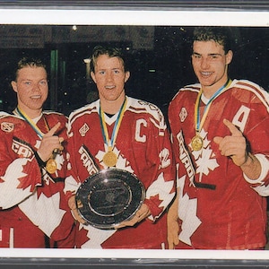 Puede incluir: Tarjeta de hockey con tres hombres con camisetas rojas con detalles blancos y la palabra "CANADA". Llevan medallas de oro y sostienen un trofeo. La tarjeta tiene un borde blanco y el logotipo de Upper Deck.