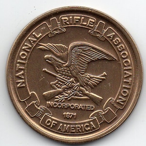 May include: A bronze coin with the National Rifle Association of America logo. The logo features an eagle holding a rifle, with the words "National Rifle Association" and "Incorporated 1871" surrounding the eagle. The coin also has the words "Of America" at the bottom.