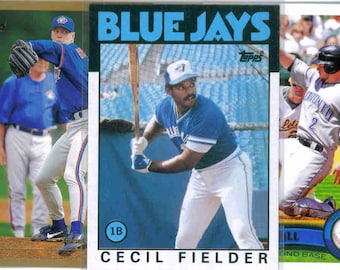 May include: A collection of baseball trading cards featuring Toronto Blue Jays players. The cards display action shots and portraits of players like Roberto Alomar, Roy Halladay, Cecil Fielder, and George Bell. The cards have the team's logo and player names.
