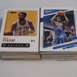 May include: Two basketball trading cards featuring New York Knicks players. The card on the left features Walt Frazier in a white and orange jersey with the text "Walt Frazier" and "Knicks". The card on the right features Julius Randle in a blue and orange jersey with the text "Julius Randle" and "Knicks".