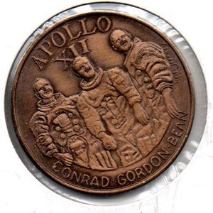 May include: A bronze-colored commemorative coin featuring three astronauts in relief. The coin is inscribed with "APOLLO XII" at the top and "CONRAD GORDON BEAN" along the bottom edge. The coin is encased in a clear plastic holder.