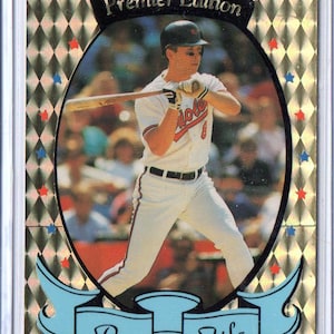 Può includere: Una carta di baseball con un giocatore di baseball in una divisa bianca e rossa con la scritta "Orioles" sul davanti. La carta ha uno sfondo dorato con un motivo a rombi e le parole "Premier Edition" e "Pro File" in nero.
