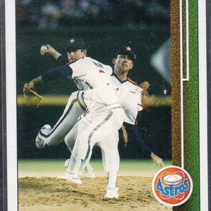 Puede incluir: Una tarjeta de béisbol con Nolan Ryan, un lanzador de los Houston Astros. La tarjeta es blanca con un borde verde y marrón. La imagen muestra a Ryan lanzando con un compañero de equipo detrás de él. La tarjeta tiene el logotipo del equipo y el nombre del jugador.